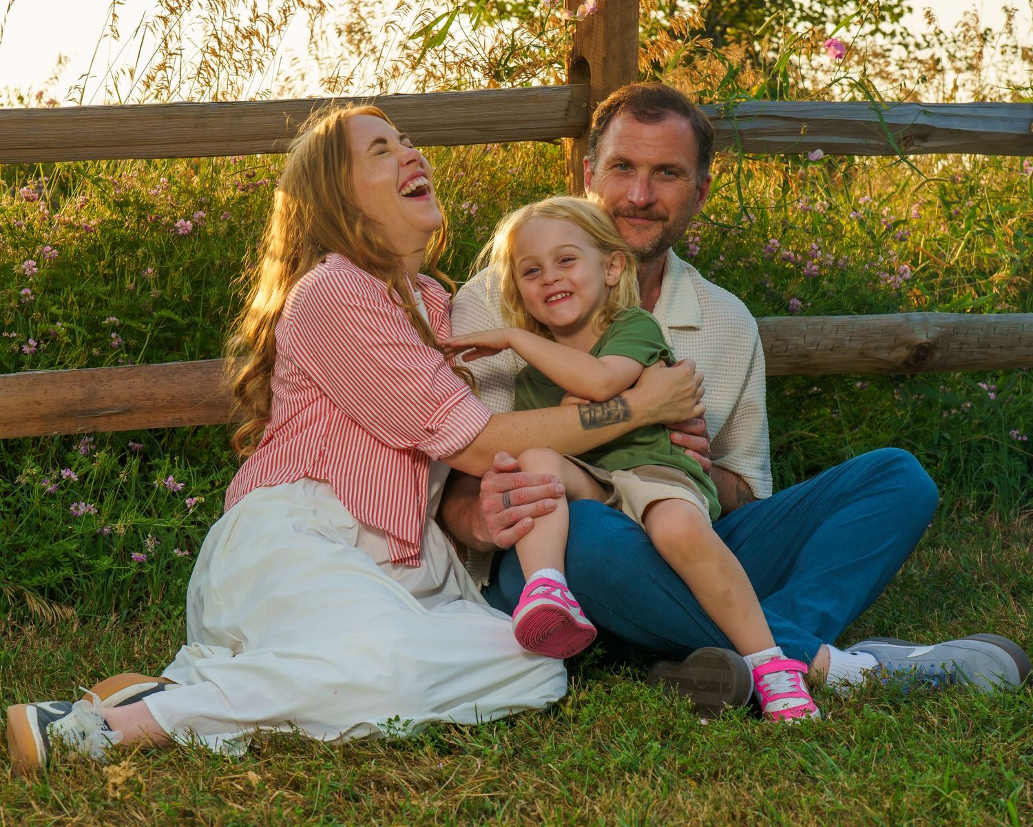 Happy family of three sitting on grass by a wooden fence, laughing and hugging at golden hour.