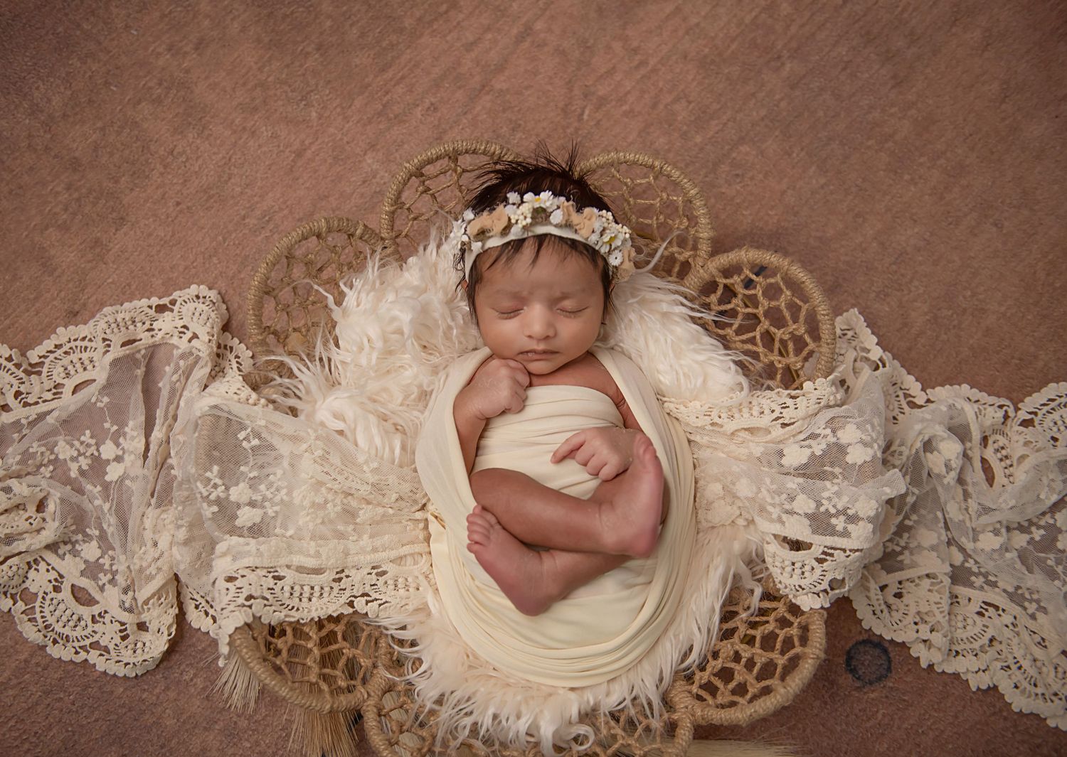 Sleeping newborn wrapped in soft cream fabric nestles on delicate vintage lace against wooden floor.