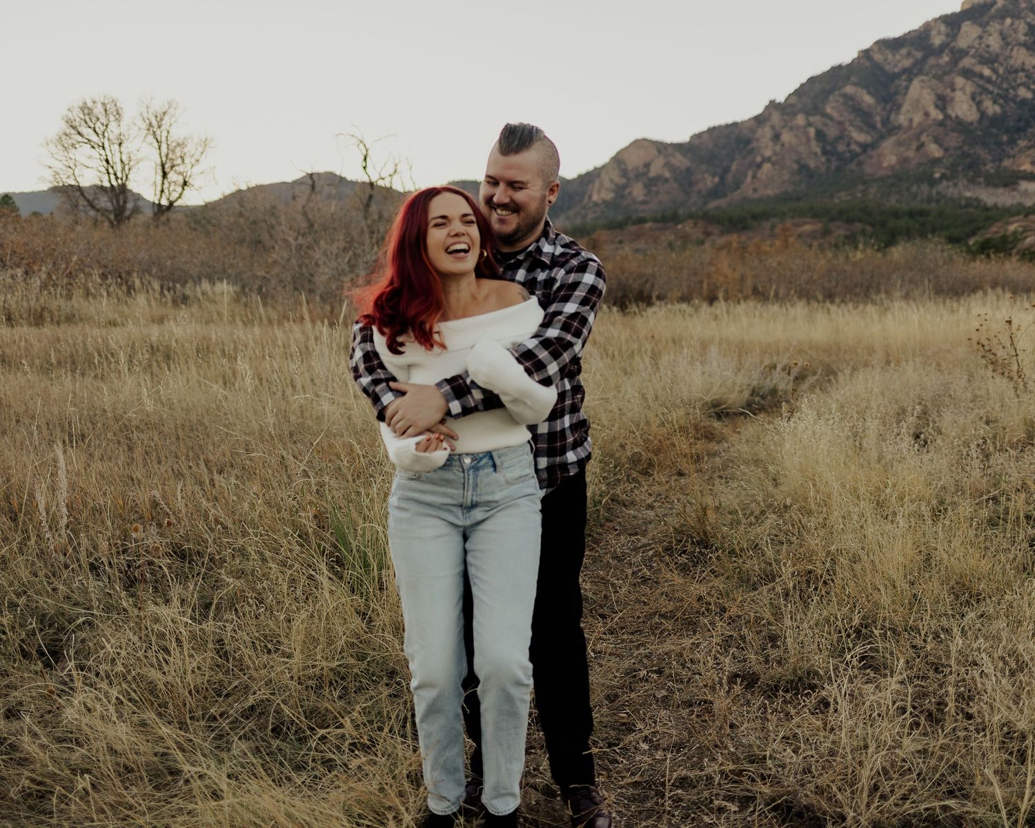 Couple shares romantic moments together in golden grass field with mountains in background during sunset.