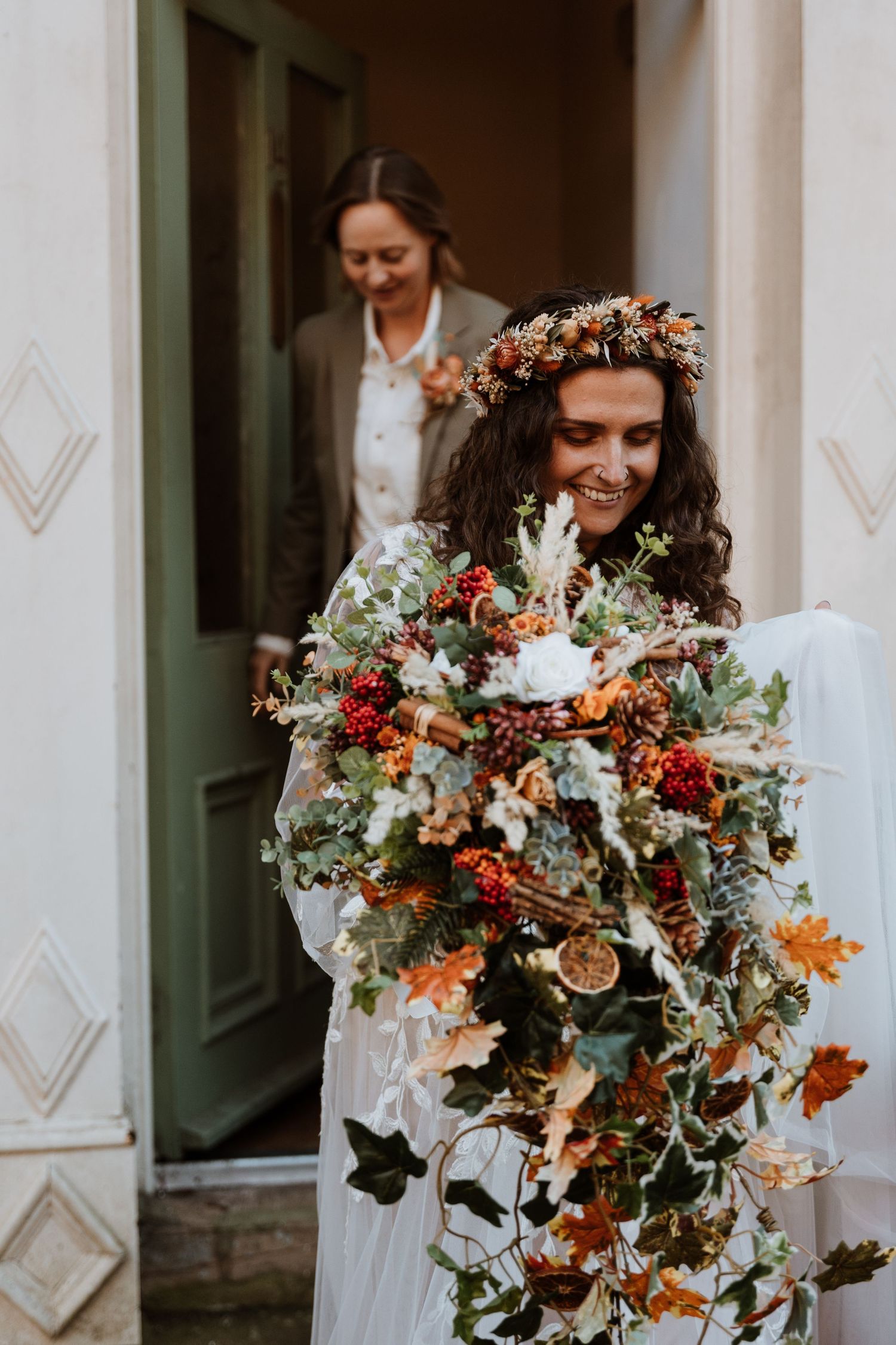 Wedding bouquet with orange and red flowers, dried elements and greenery in fall tones being held in front of white walls.