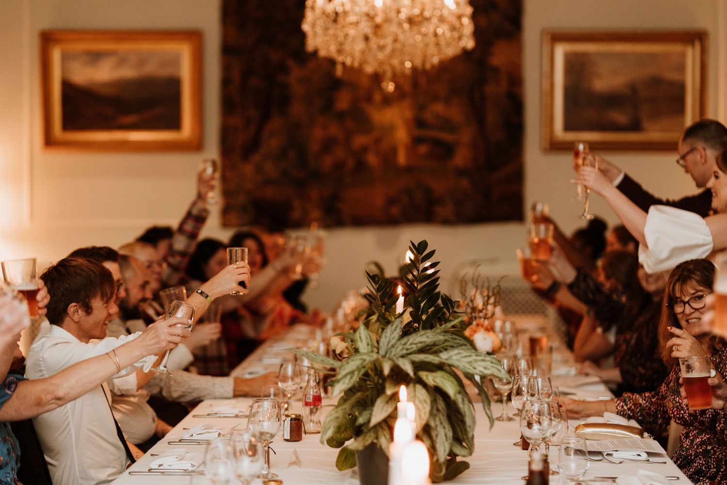 Dinner guests raise their glasses for a toast at an elegant table decorated with greenery under a crystal chandelier.