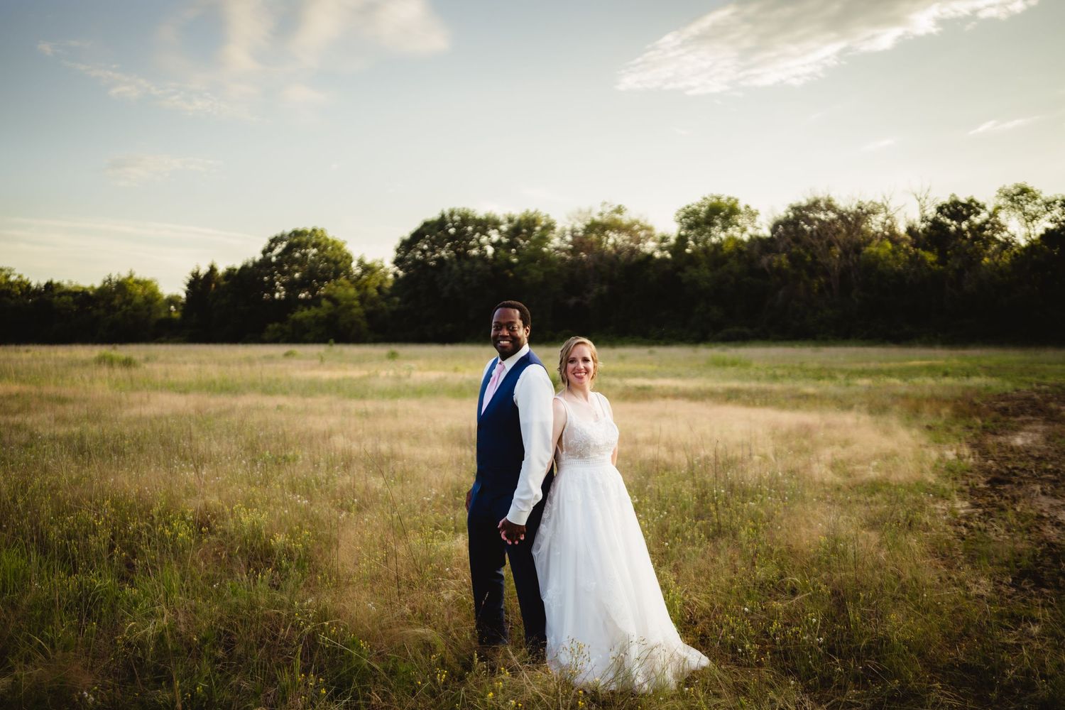 A couple stands together in a golden meadow at sunset wearing formal wedding attire.