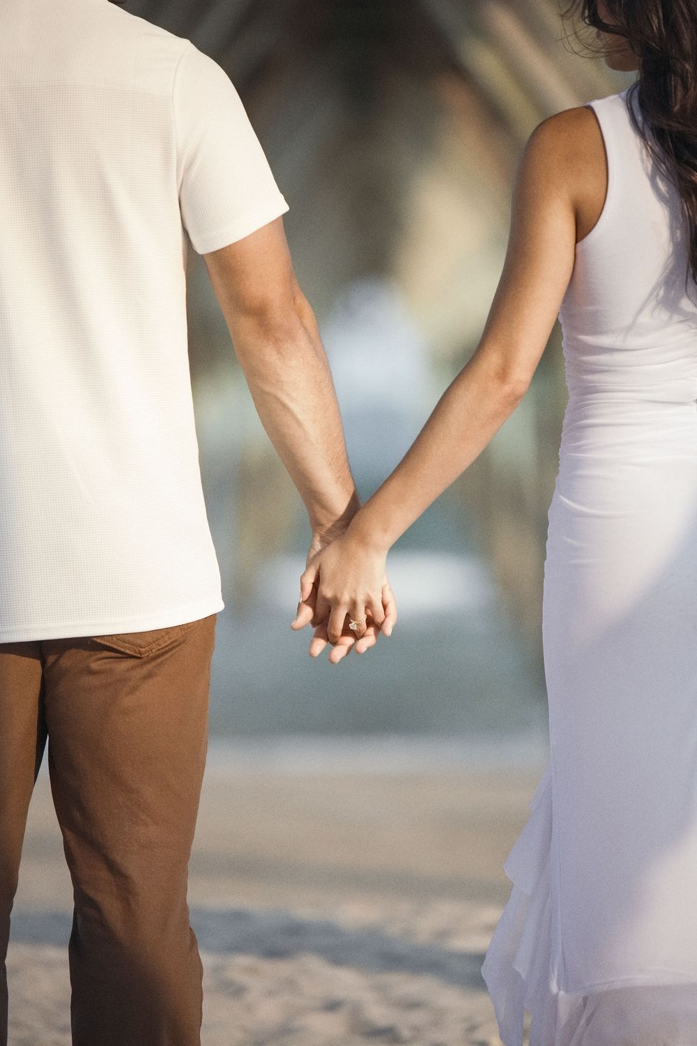 Close-up of couple holding hands on sandy beach wearing white and brown outfits.