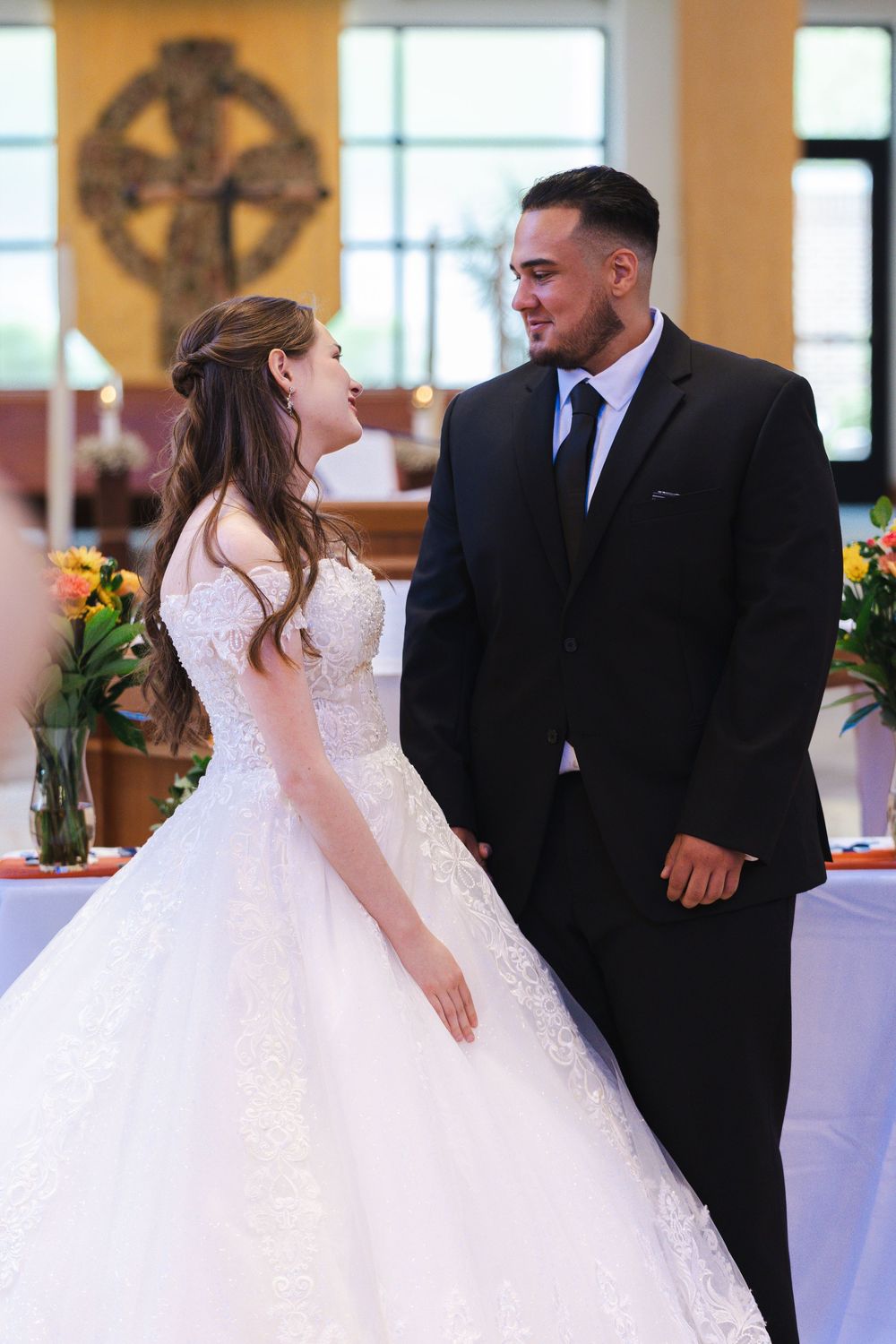 A bride in a white lace gown and groom in black suit share a romantic moment during their church wedding ceremony.
