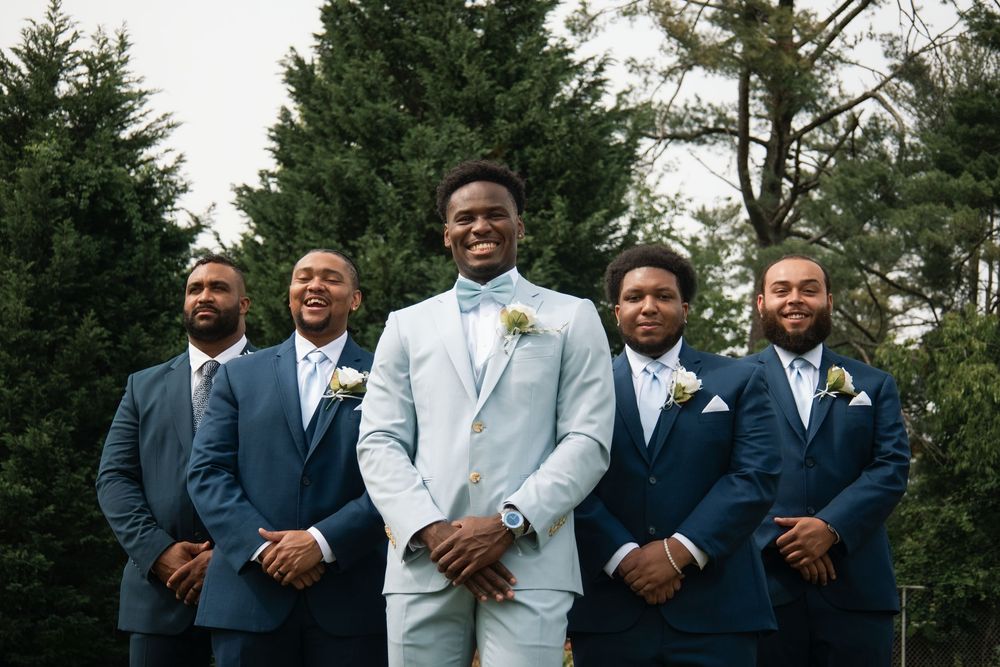 Groomsmen in navy suits and one in white pose together outdoors by evergreen trees.