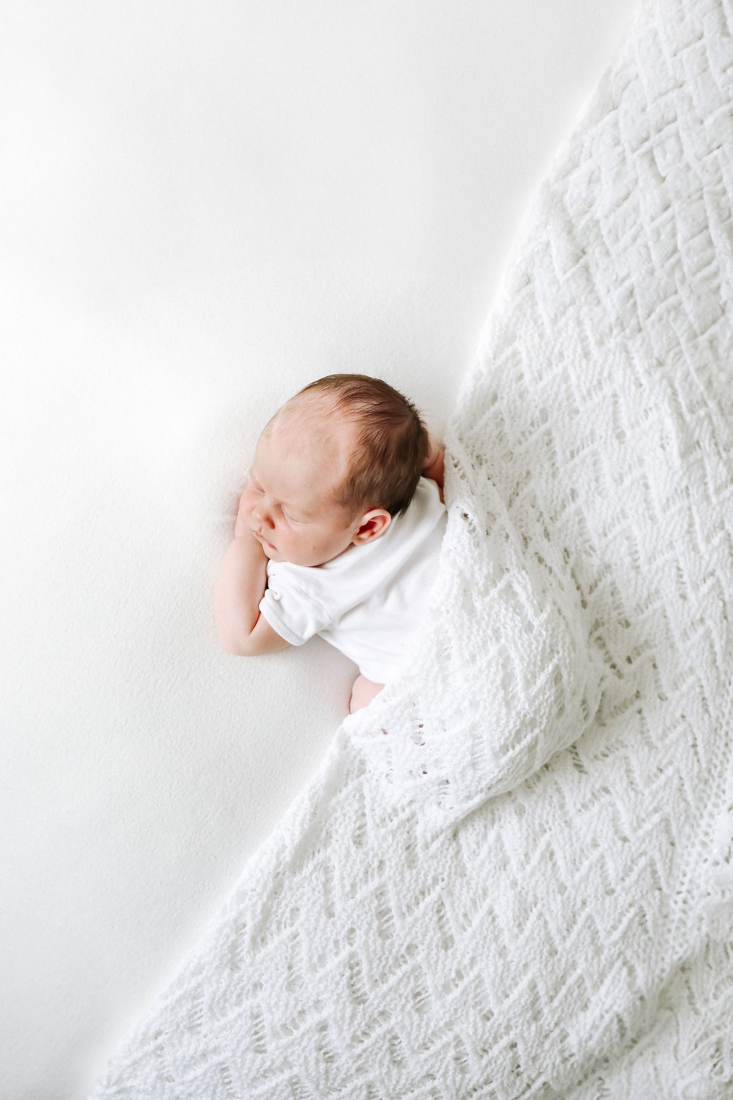 Sleeping baby wrapped in white blanket lying on textured white bedding in minimalist style, during newborn photoshoot in Nottingham