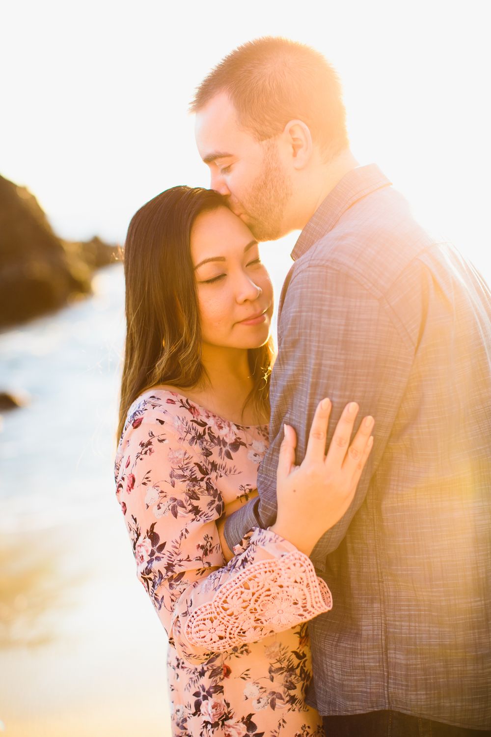 Romantic backlit engagement photos of couple embracing at sunset on beach in soft golden light.