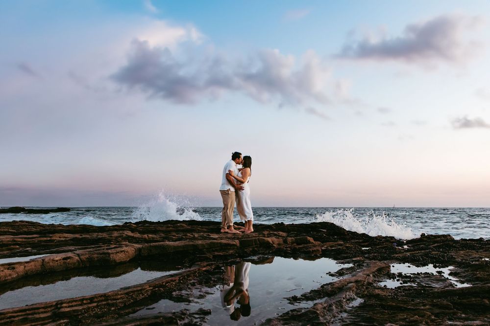 A couple embraces on rocky cliffs overlooking dramatic ocean waves crashing at sunset during engagement session in Newport Beach.