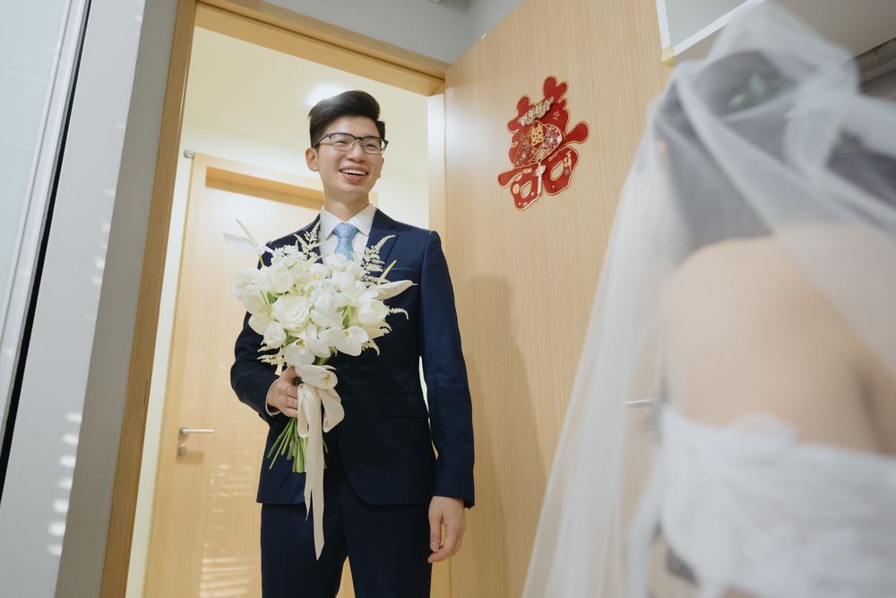 Traditional Chinese wedding ceremony featuring a groom in a dark suit holding a white lily bouquet by a doorway with red decor.