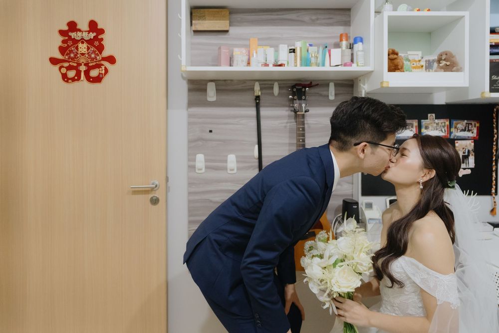 A pair in wedding attire share a tender kiss in front of built-in shelves displaying medicine bottles and supplies.