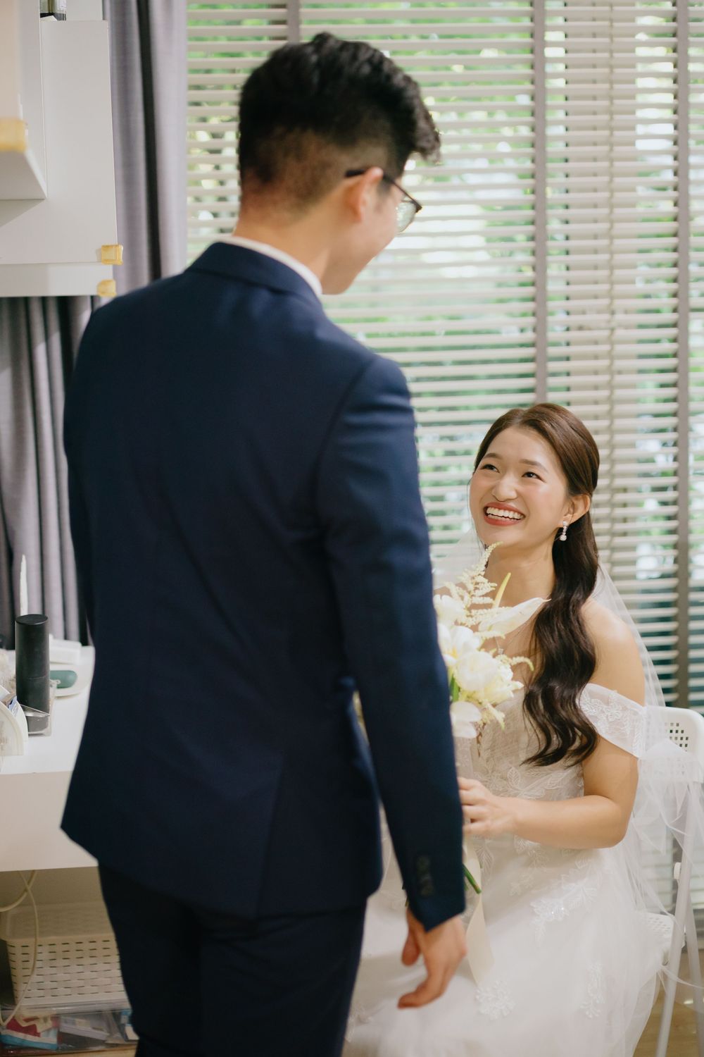 Bride in white dress and groom in navy suit share a joyful moment by a window with venetian blinds.