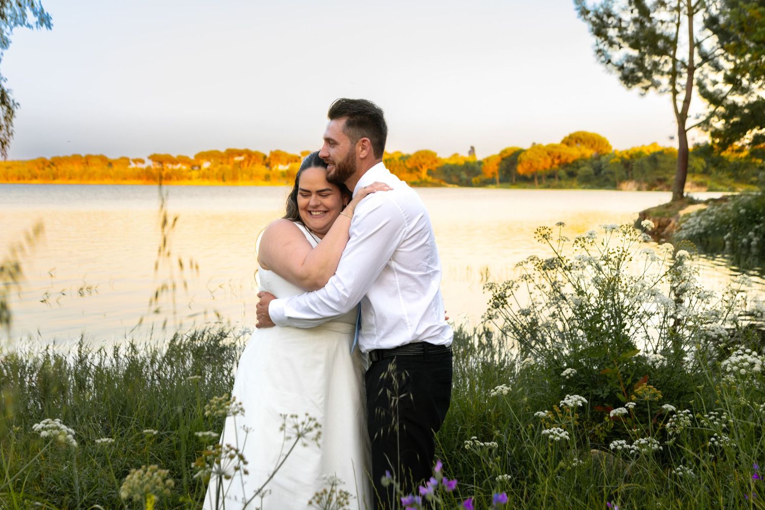Couple embracing in field of wildflowers during golden sunset by the water.