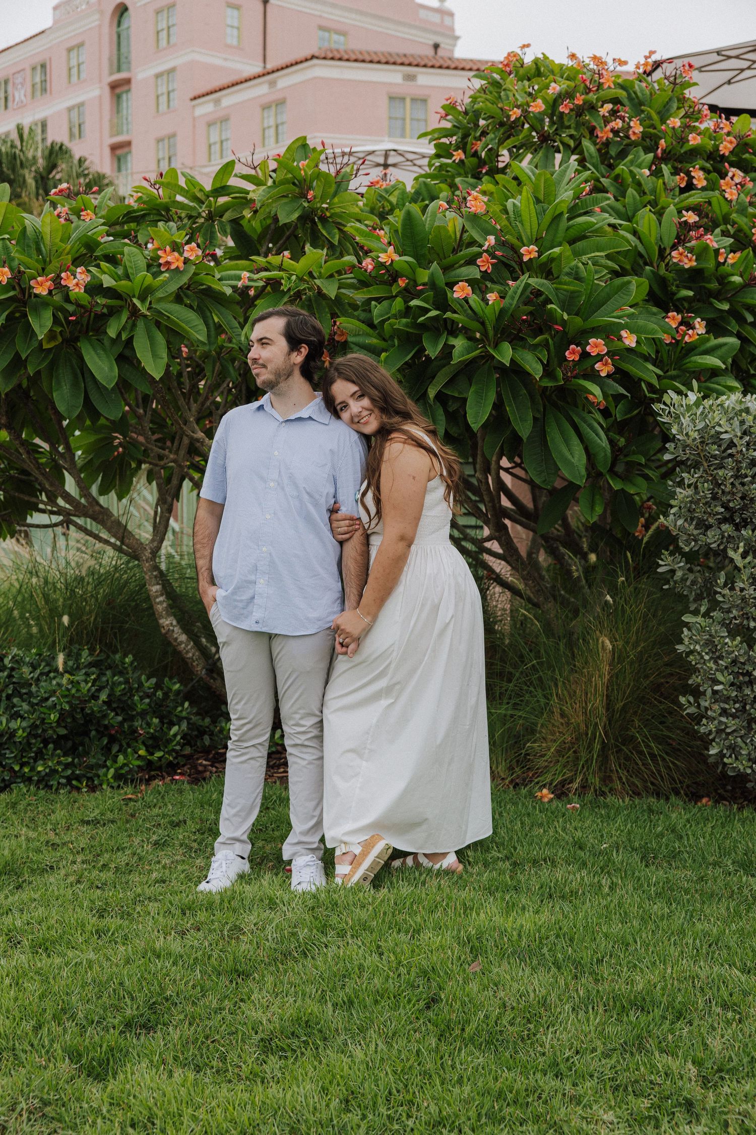 A couple stands together on a lush green lawn with flowering plumeria trees in the background.