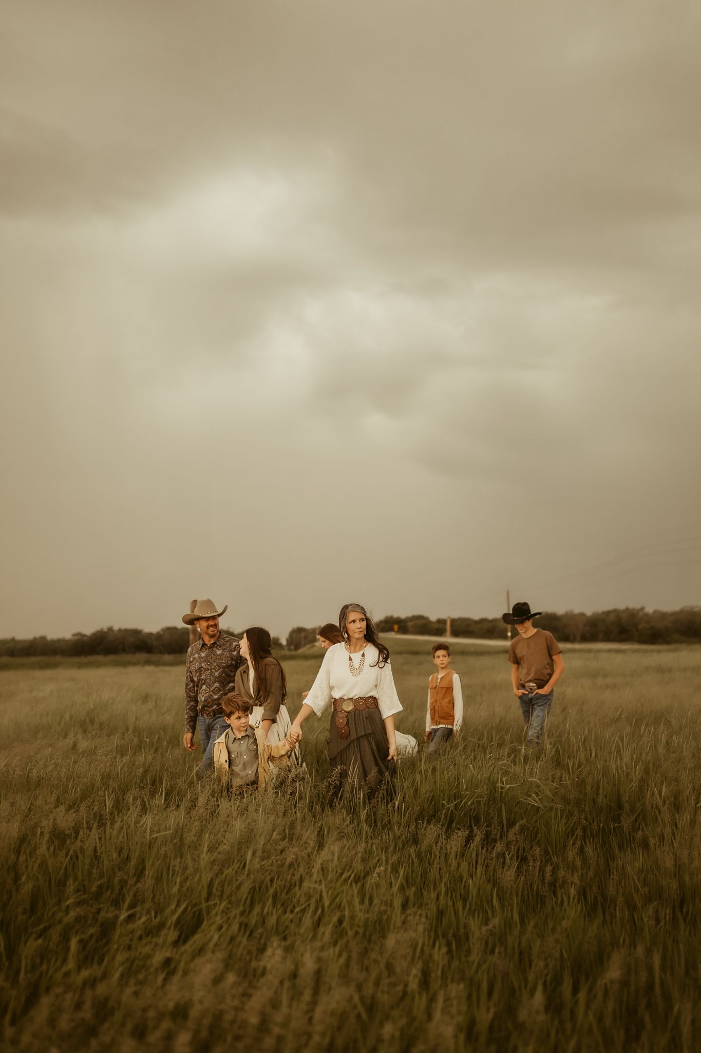 A group in casual attire sits together in a tall grass field as clouds loom overhead in a sepia-toned photograph.