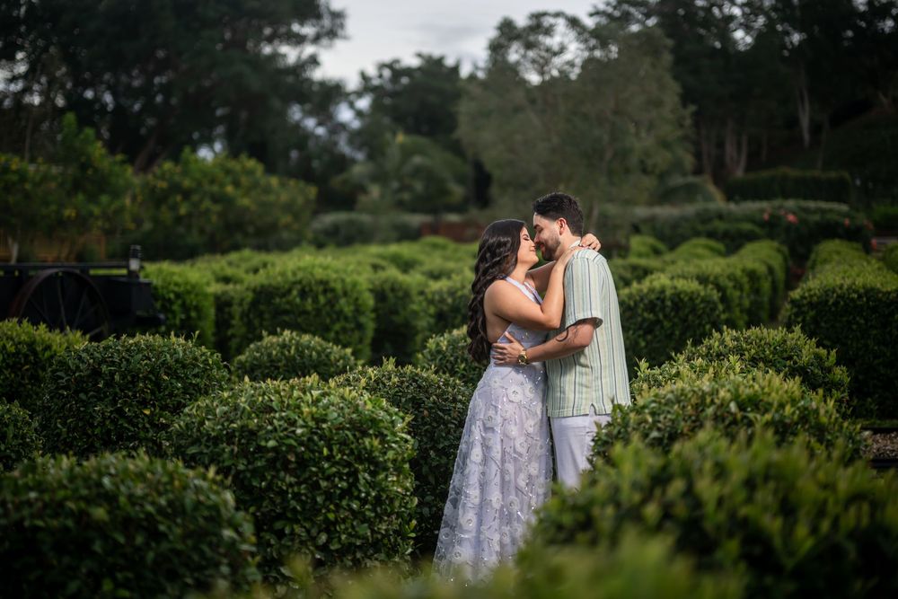 Two people share an intimate embrace among manicured hedges in a lush garden setting at dusk.