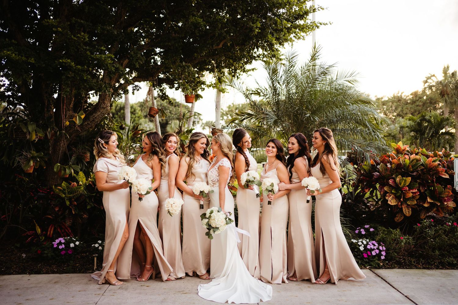 A bridal party poses together in champagne colored dresses amid lush garden scenery at sunset.