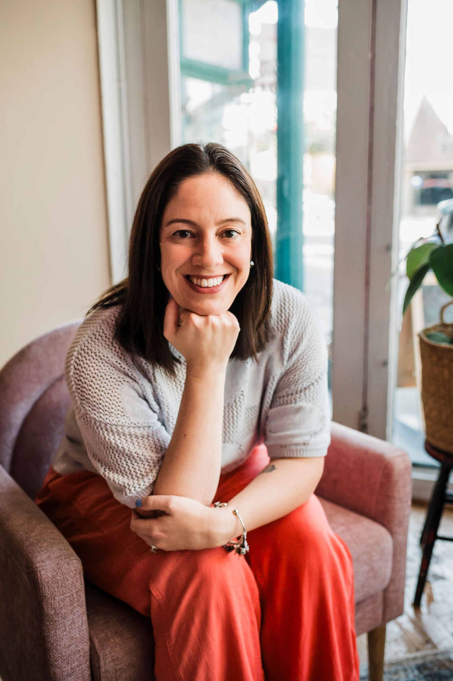 Person wearing grey sweater and coral skirt sits comfortably in upholstered armchair by window with natural light streaming in.