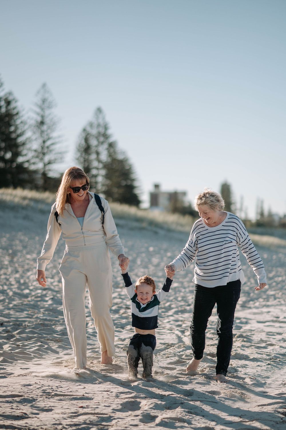 Three generations walk together on a sandy beach at sunset with evergreen trees in the background.