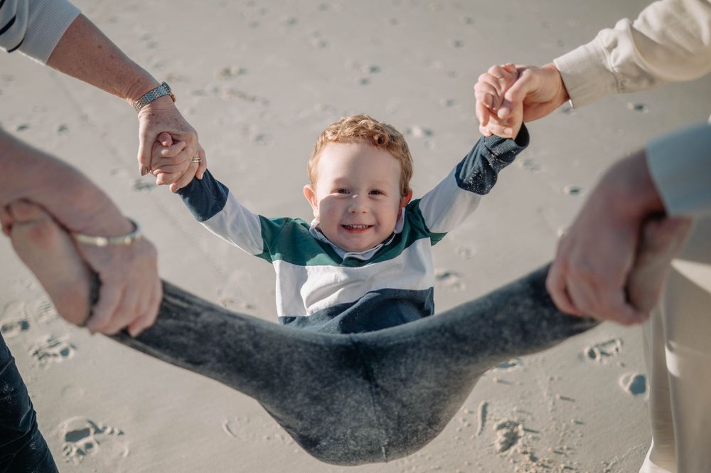 Child being swung by both hands while smiling and wearing a striped shirt on sandy ground.