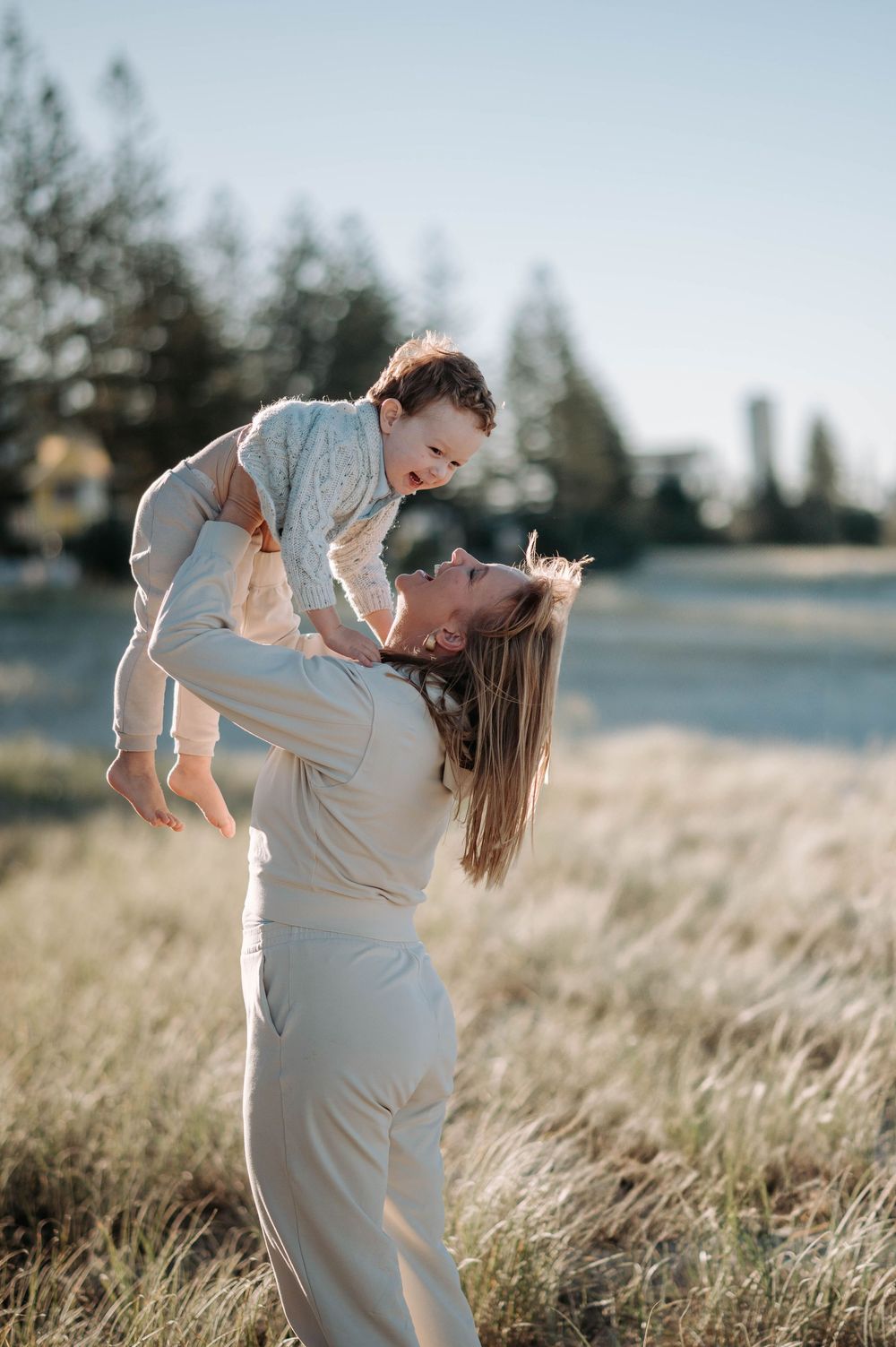 Parent and child share a joyful moment outdoors in a sunlit field with trees in the background.