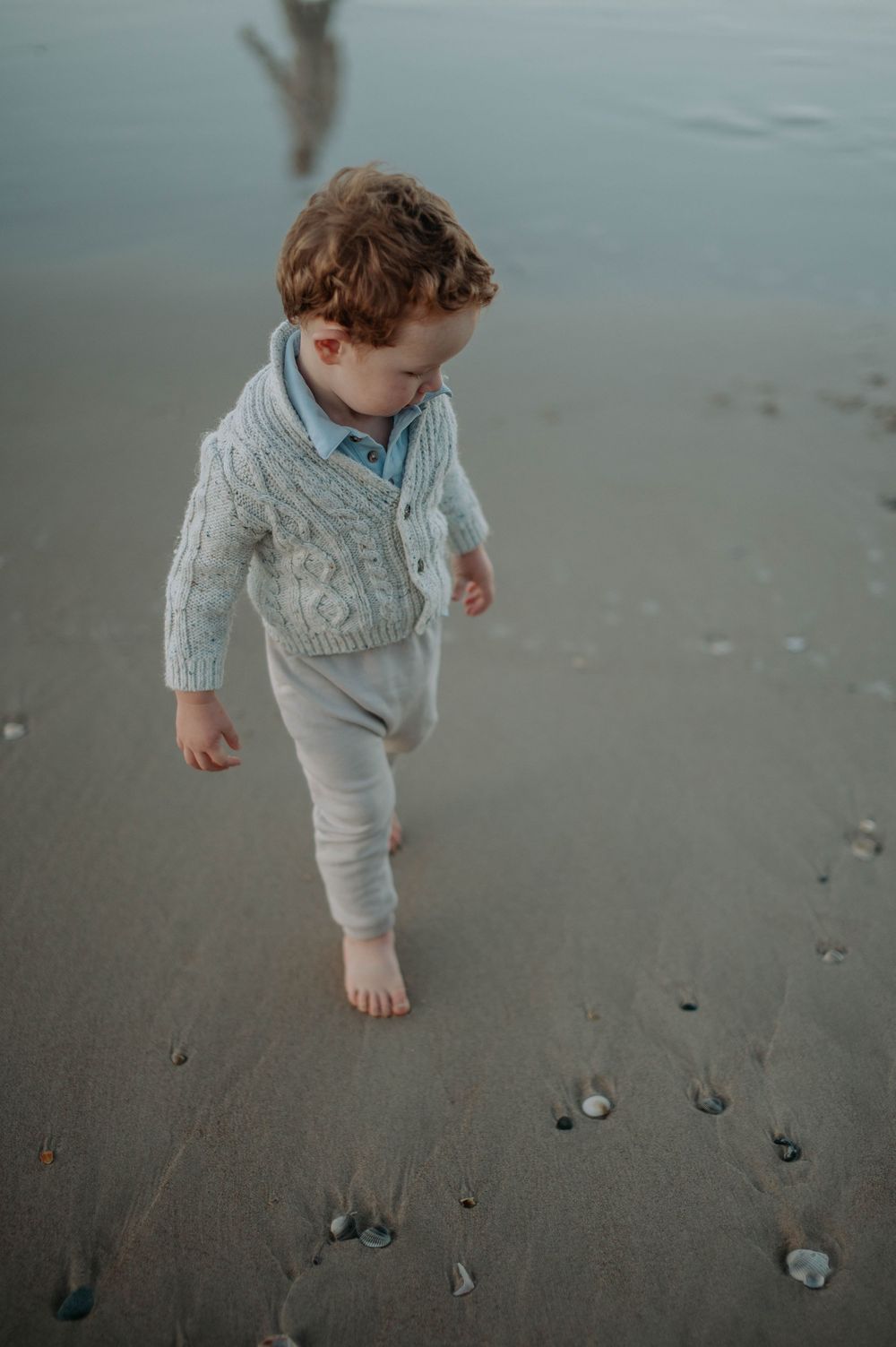 Small child in cozy sweater walking barefoot on wet sand at the beach.