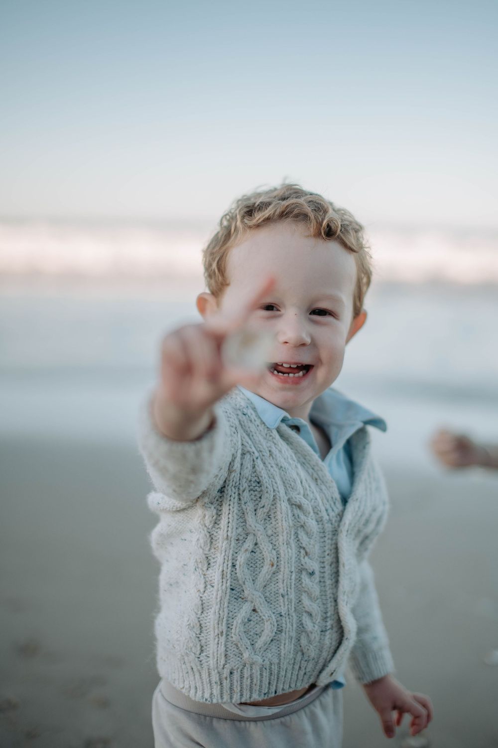 A joyful toddler in a knit sweater plays and waves at the beach during sunset.