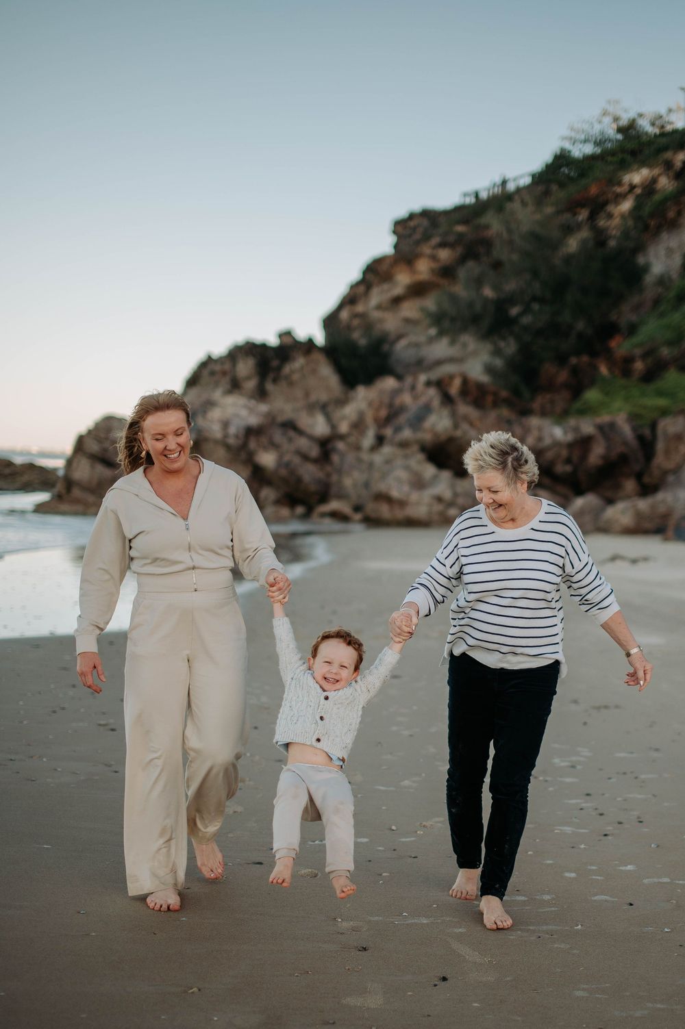 Three generations enjoying a playful walk together on a sandy beach with rocky cliffs.