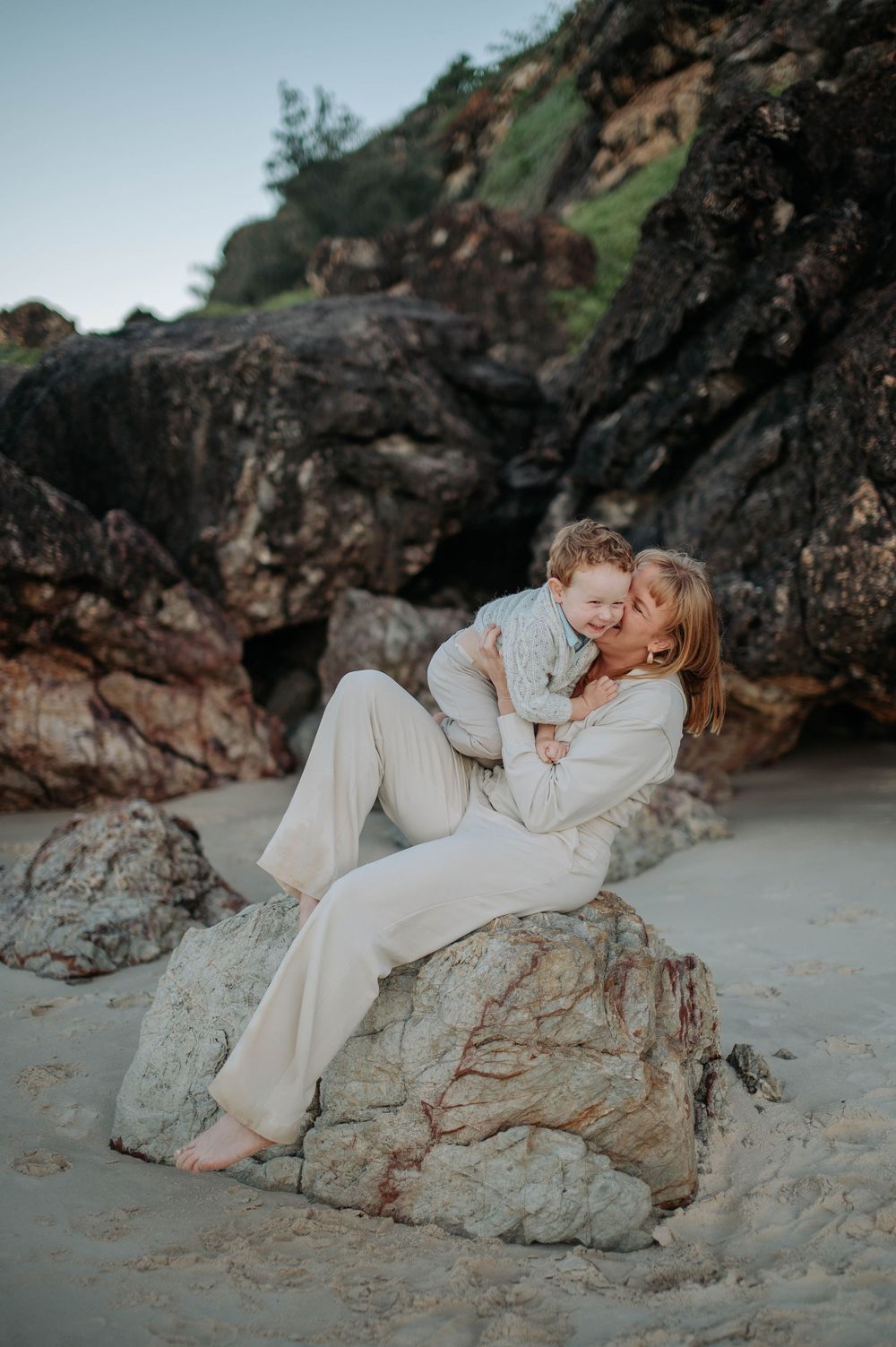 A tender beach photoshoot moment with woman in white sitting on coastal rocks at sunset.