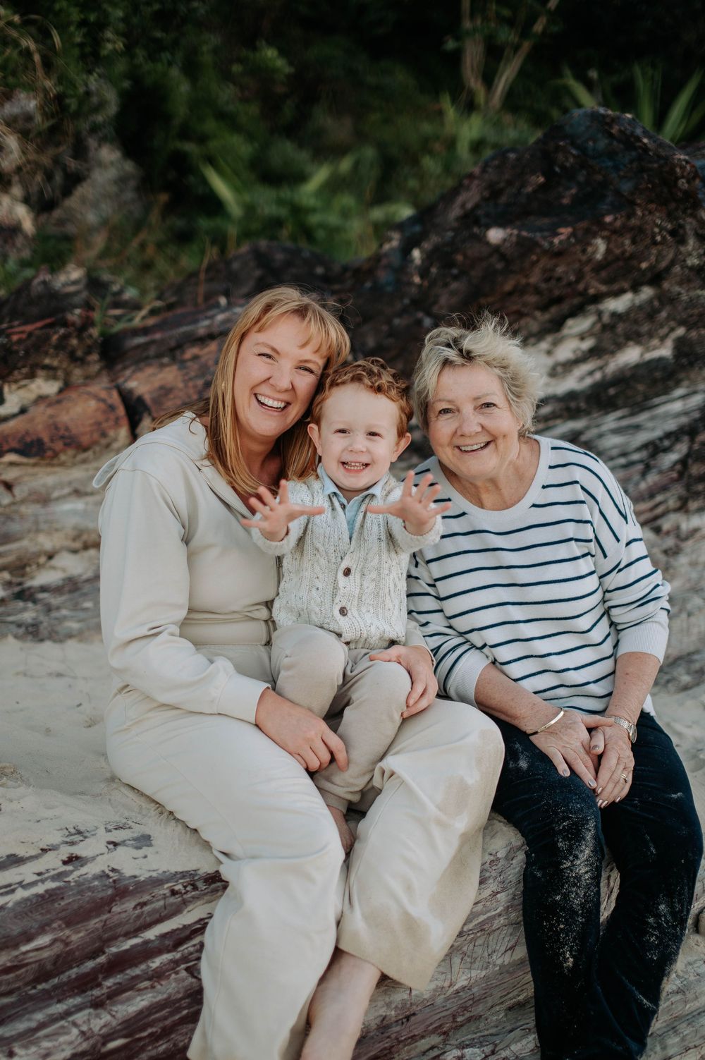 Three generations share a joyful moment together while sitting on driftwood at the beach.