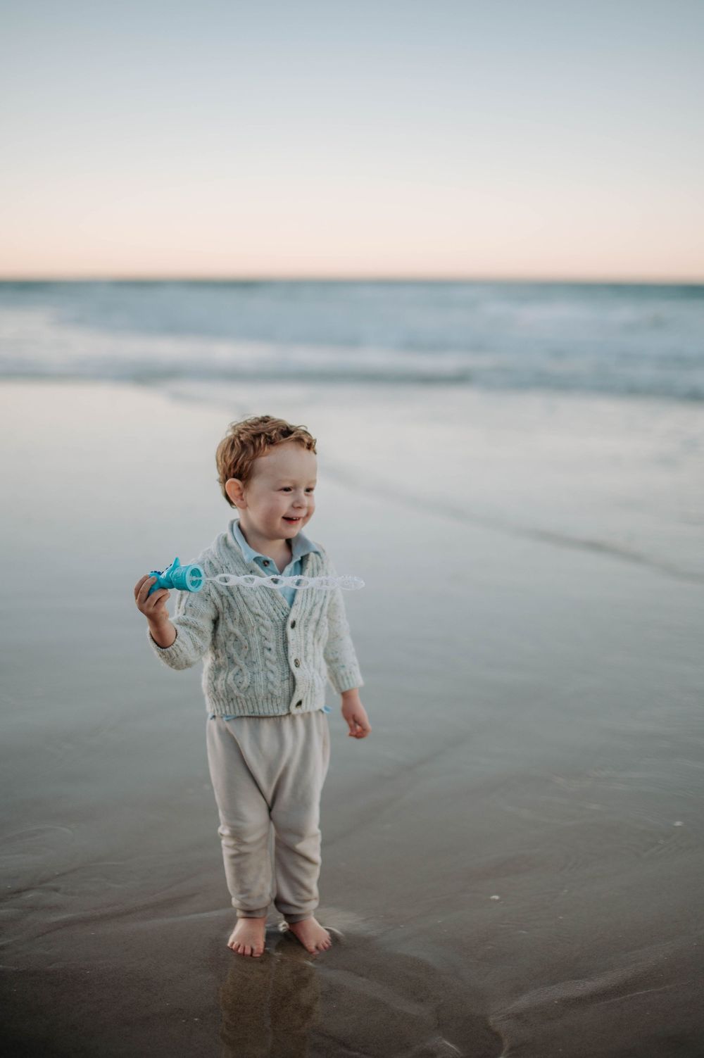 Joyful toddler playing barefoot on a serene beach at dusk with calm ocean waters in the background.