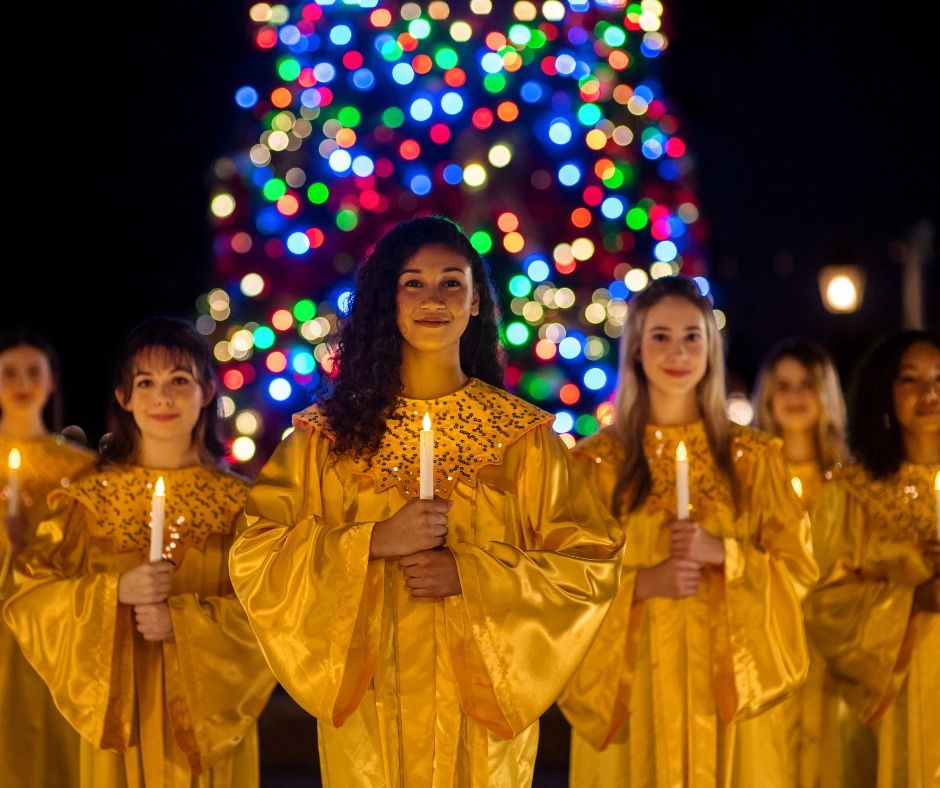 A choir in golden robes holds candles in front of a colorfully lit Christmas tree.
