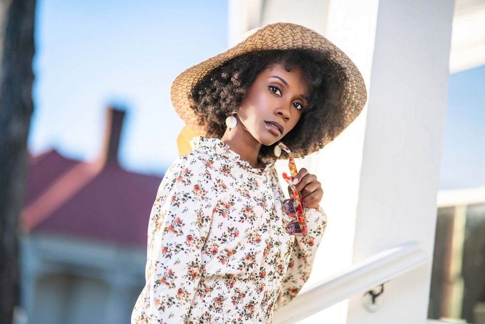 Person in a floral dress and sun hat posing against a white wall outdoors.