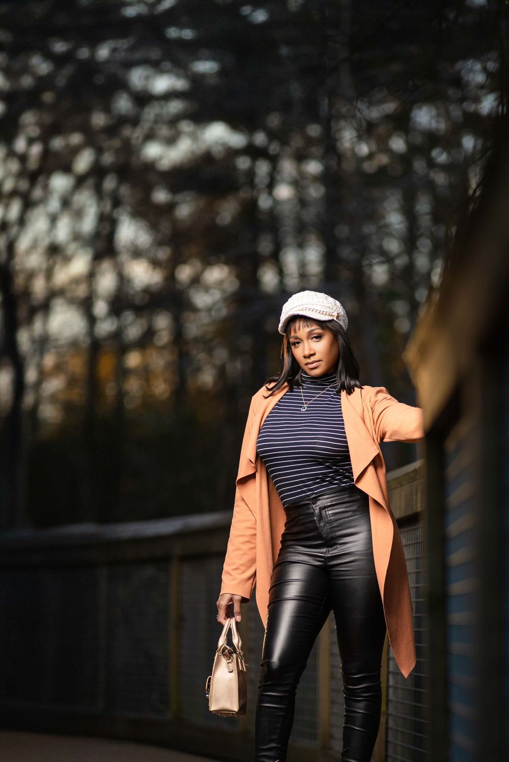 Stylish fall fashion featuring a striped turtleneck, leather pants and peach coat against a moody forest backdrop.