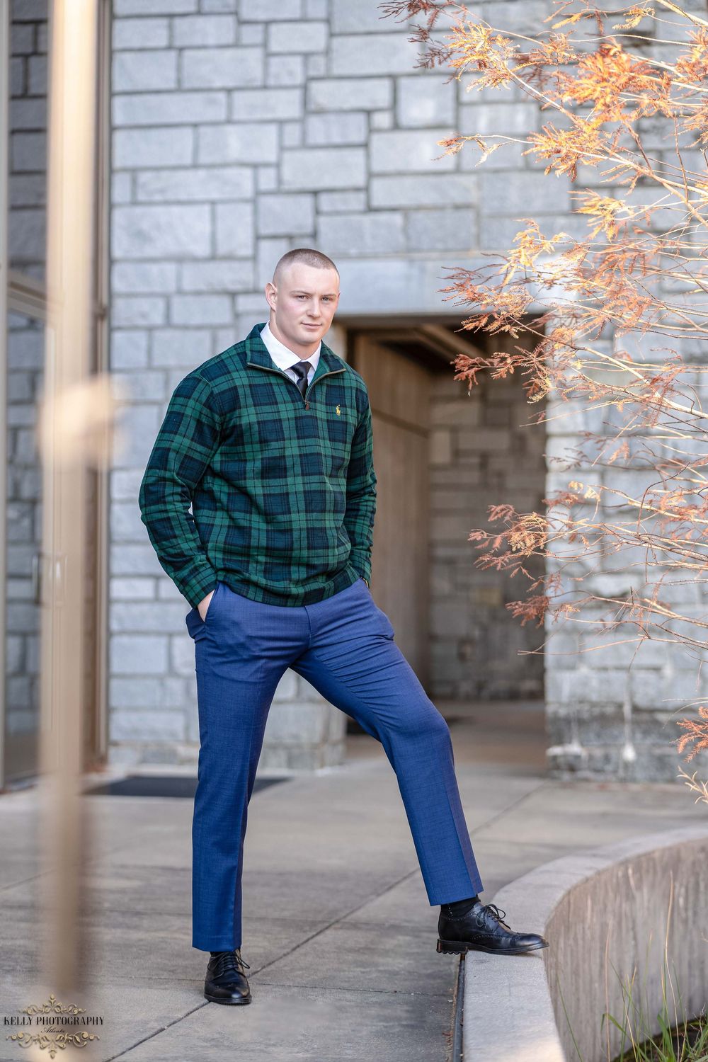 Man in plaid green sweater and blue pants poses confidently against stone wall with autumn foliage.
