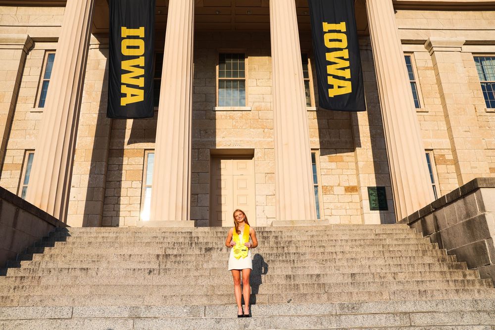 Graduate in black cap and gown poses on stone steps near classical columns with yellow banners at sunset.