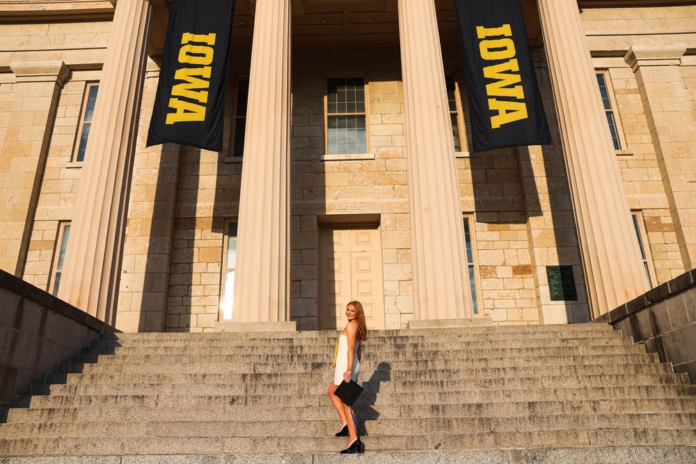 Wide view of graduate posing on steps of historic stone building with yellow university banners.