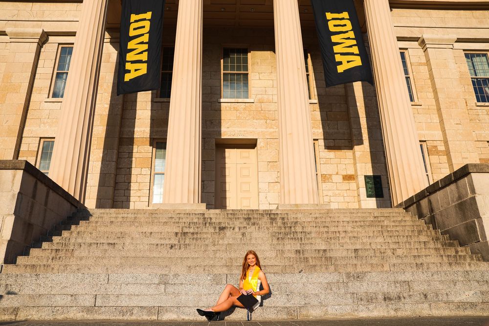 Graduate sits on steps of University of Iowa building with yellow IOWA banners in warm sunlight.