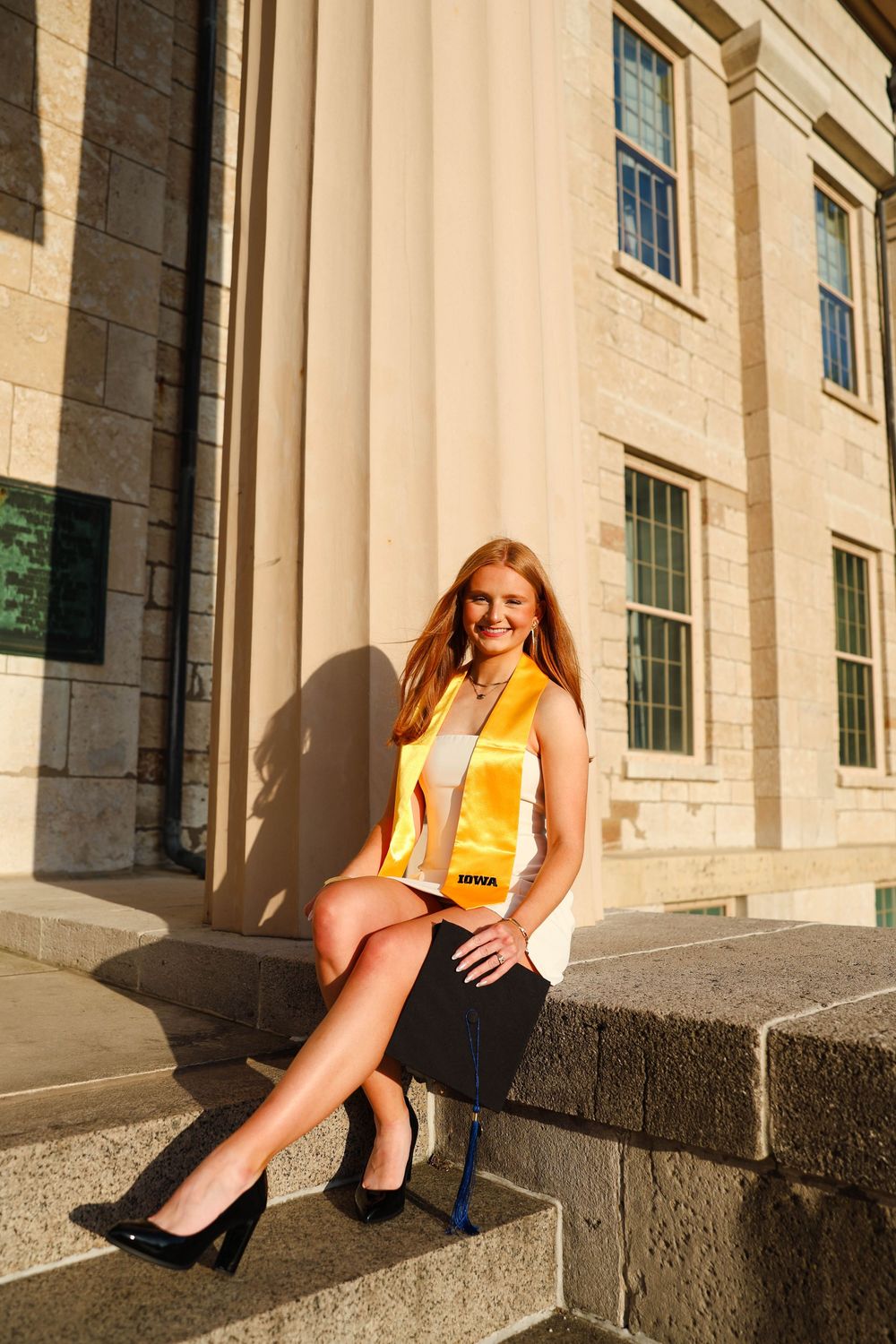 Graduate poses on stone steps wearing yellow and white outfit with black heels at University of Iowa.
