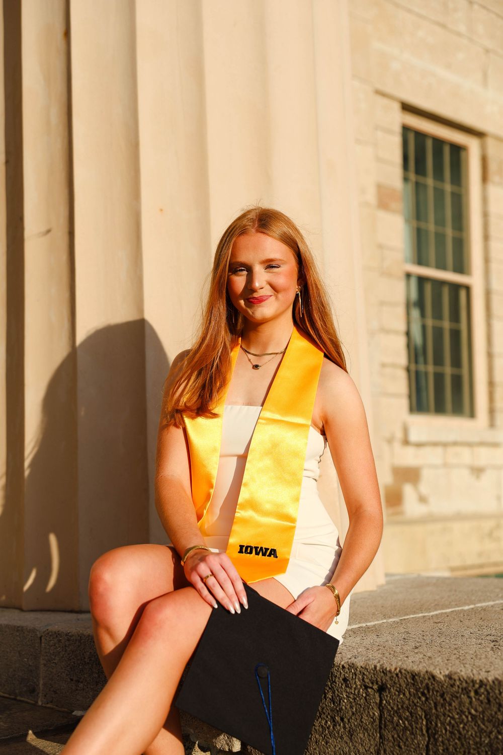Graduate wearing yellow stole sits on stone ledge near classical columns in warm sunlight.