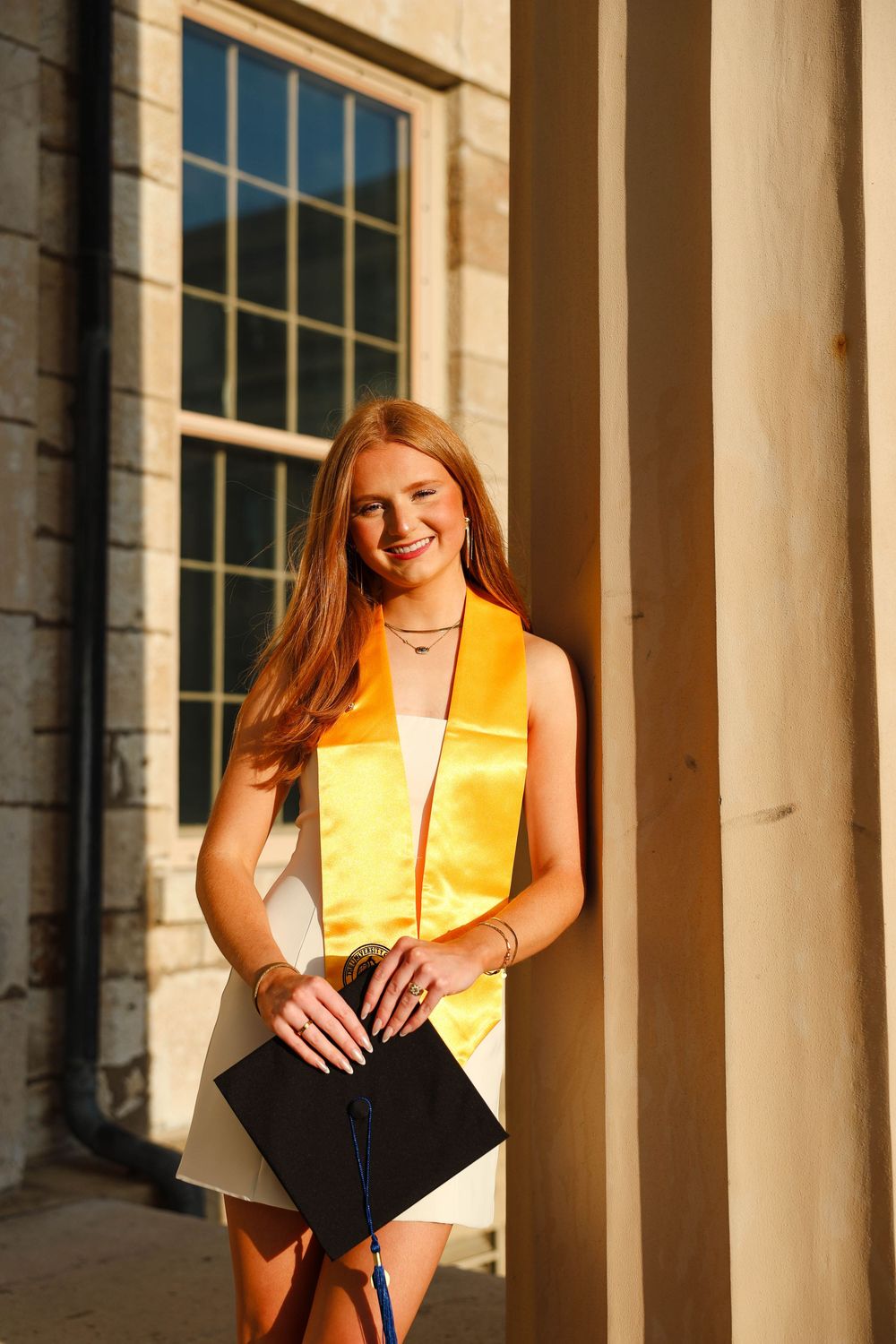 Graduate wearing black cap and gown with gold stole poses in front of classical stone columns during sunset.