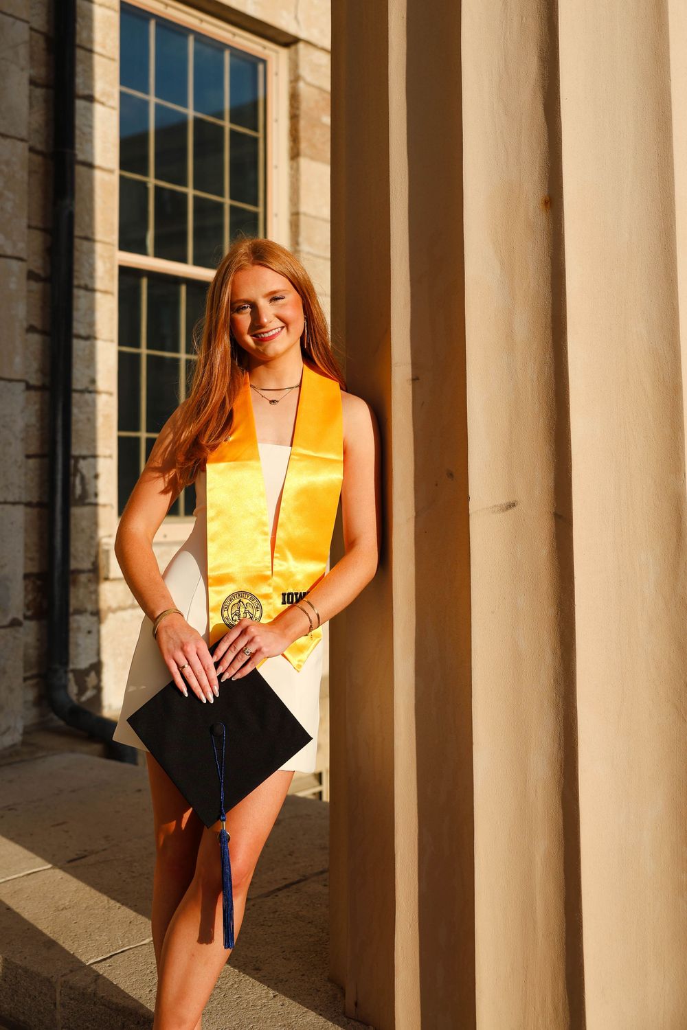 Graduate in yellow stole poses among stone columns of university building at golden hour.
