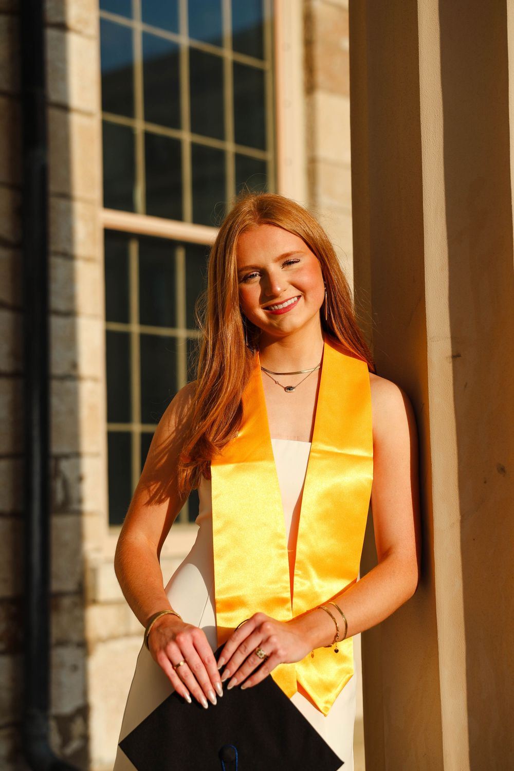 Graduate wearing yellow honor stole poses against stone columns in graduation photo series.