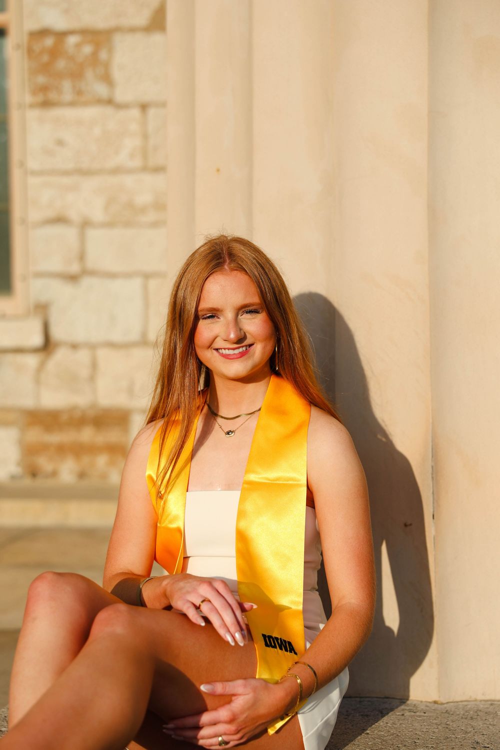 Graduate sits on stone ledge in golden hour light wearing graduation stole and holding diploma.