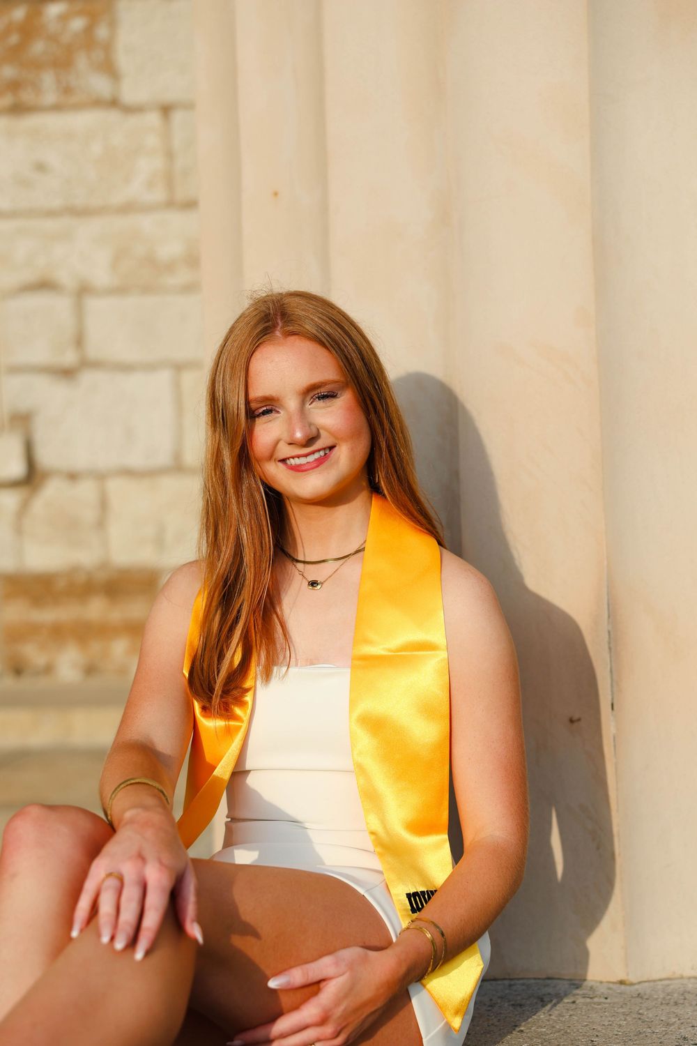 Graduate in white dress and gold sash poses against stone wall in sunlight at graduation.