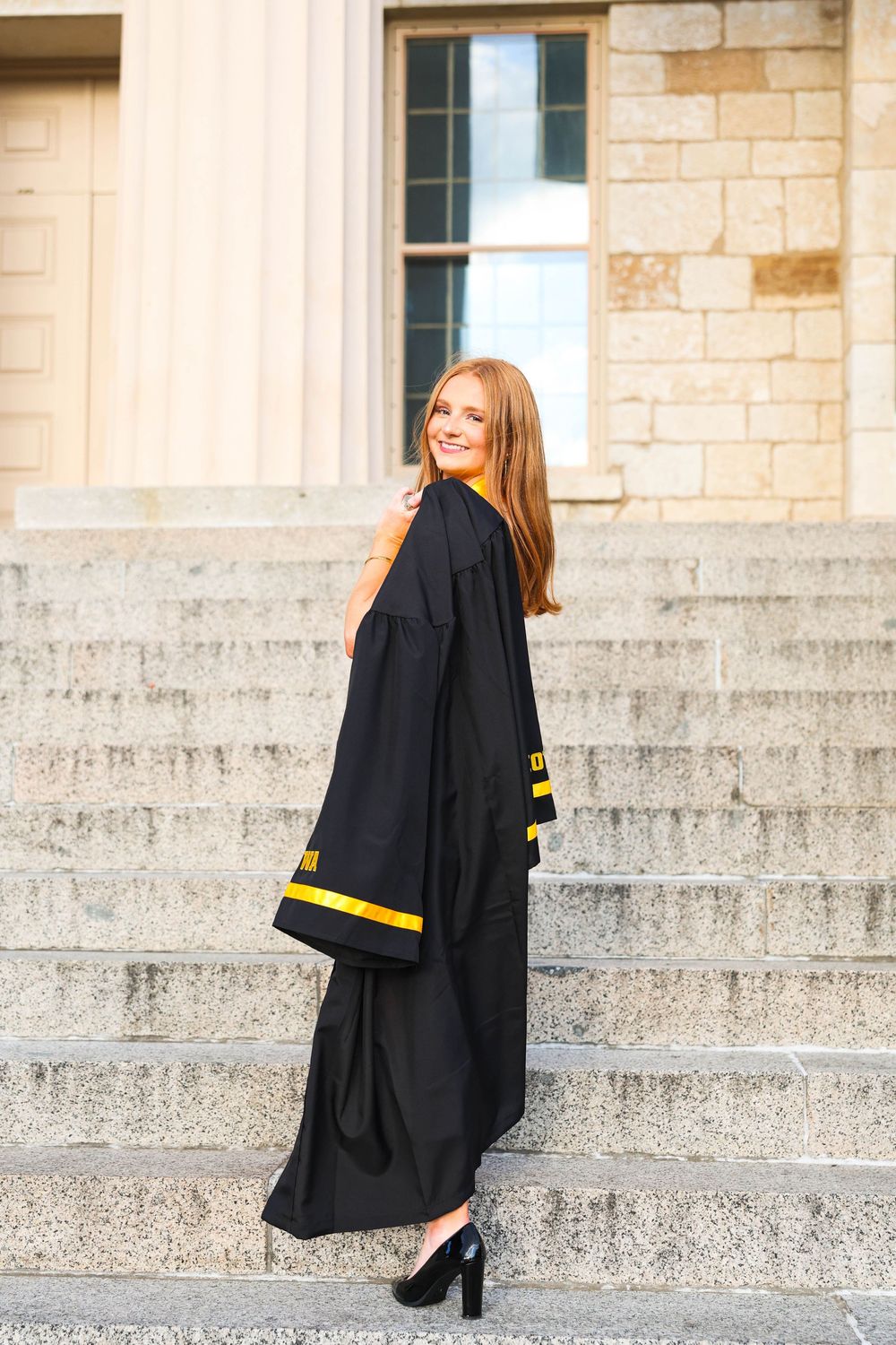 Graduate in black gown with gold trim poses on stone steps for professional graduation photos.