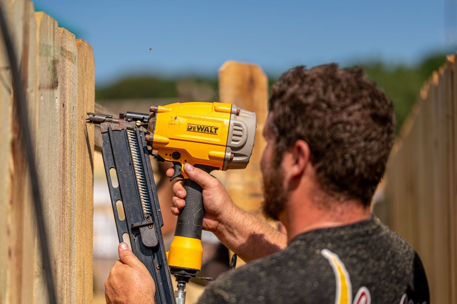 Someone using a yellow DeWalt nail gun to attach boards while building a wooden fence outdoors.
