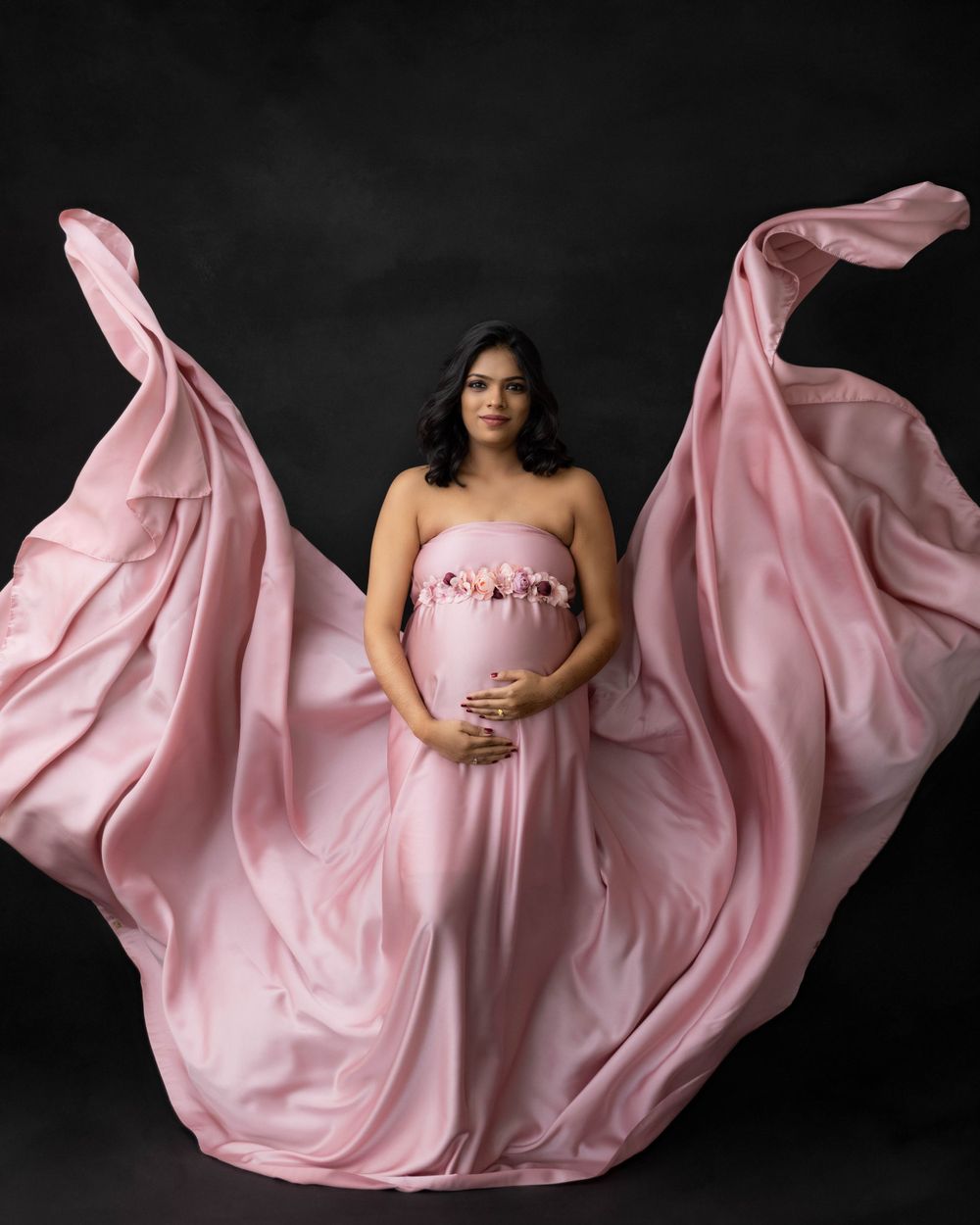 Dramatic maternity portrait with floating pink fabric creating wing effect against dark background.