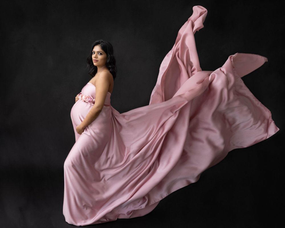 Ethereal maternity photo with flowing pink dress fabric creating movement against black backdrop.