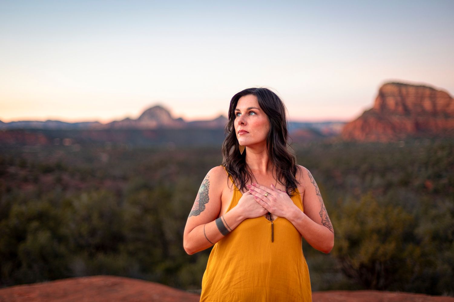 Woman in yellow dress with tattoos meditating at sunset with Sedona red rocks behind her.