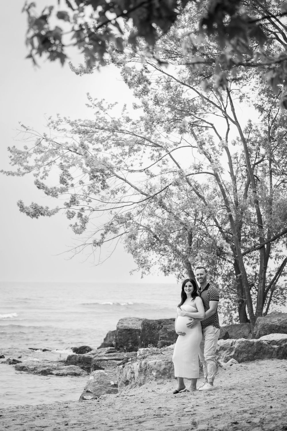 Romantic black and white beach photograph showing a couple embracing beneath a tree on a rocky coastline.